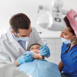 A dentist performs a checkup on a child, assisted by a dental nurse positioning an overhead light