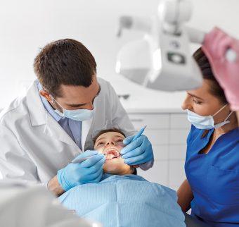 A dentist performs a checkup on a child, assisted by a dental nurse positioning an overhead light