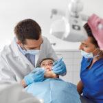 A dentist performs a routine checkup on a child, as a nurse looks on.