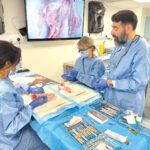 Dr Ali explains a proceedure to two students sitting at a table covered with dental equipment.