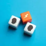 One orange and two white cubes on a flat blue background, the cubes symbolise conflict with two human silhouettes on the white cubes and a lightening strike on the orange.