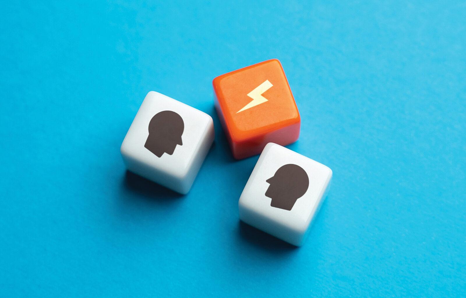 One orange and two white cubes on a flat blue background, the cubes symbolise conflict with two human silhouettes on the white cubes and a lightening strike on the orange.