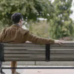 A man in a brown suit sits on a park bench by himself, facing away from the camera, he has his right arm stretched out across the top of the otherwise empty bench.