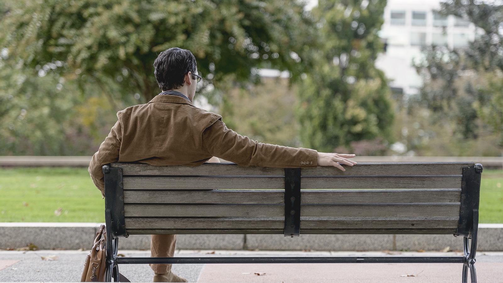 A man in a brown suit sits on a park bench by himself, facing away from the camera, he has his right arm stretched out across the top of the otherwise empty bench.